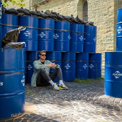 Porträtfoto von Stefan Woischner mit Sonnenbrille, sitzend vor gestapelten blauen FUCHS-Fässern auf Kopfsteinpflaster