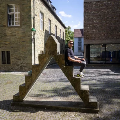 Junger Mann mit Sonnenbrille sitzt seitlich auf einer steinernen Skulptur mit Treppenform in einem Innenhof, Reise- und Porträtfotografie