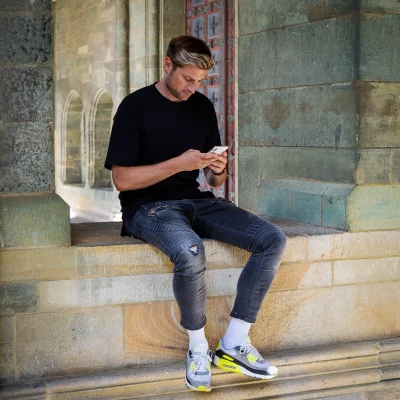 Fotoshooting mit Stefan Woischner: Junger Mann mit Smartphone, lässig sitzend auf einem Steinsims im Außenbereich, leger gekleidet in schwarzem T-Shirt und Jeans