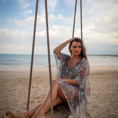 Frau in lockerem, gemustertem Kleid sitzt auf einer Holzschaukel am Strand mit Meer im Hintergrund
