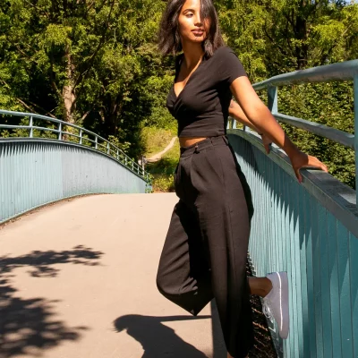 Weibliches Model in schwarzem Outfit posiert lächelnd auf einer Brücke bei Tageslicht, umgeben von grünen Bäumen