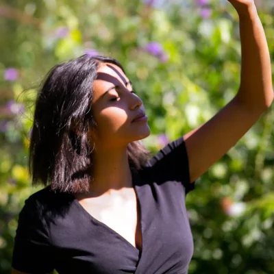 Frau mit kurzen dunklen Haaren, die in schwarzem Oberteil im Sonnenlicht steht und die Hand schützend vor das Gesicht hält, grüner Naturhintergrund
