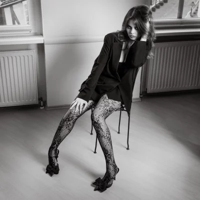 Junge Frau in einem schwarzen Blazer und einer gemusterten Strumpfhose, die auf einem Stuhl sitzt.