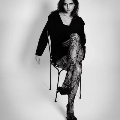 Schwarzweiss Fotoshooting; Frau mit überschlagenen Beinen auf einem Stuhl in einem Fotostudio, trägt gemusterte Netzstrumpfhose und Jacke.