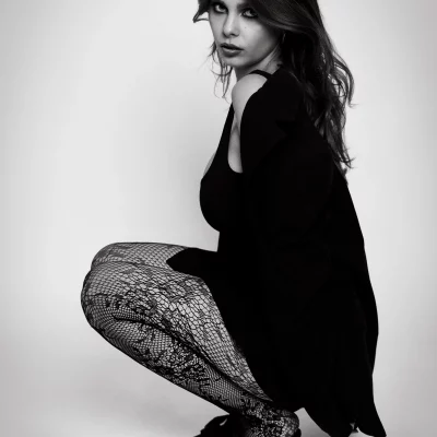 Frau mit langen Haaren in Netzstrumpfhose und hohen Schuhen hockt seitlich vor Studio Hintergrund