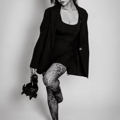 Fashion Schwarzweiß Fotoshooting: Model Valeriia in schwarzem Kleid und gemusterten Netzstrümpfen hält High Heels in der Hand und steht auf einem Bein