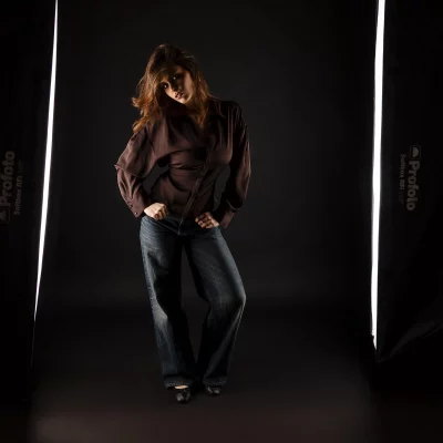 Frau in dunkler Bluse und Jeans steht mit verschränkten Armen und leicht geneigtem Oberkörper zwischen zwei vertikalen Lichtstreifen im Fotostudio