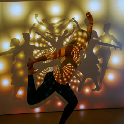 Porträtfotografie eines weiblichen Models in Tanzpose, beleuchtet von Projektionen abstrakter Lichter und Silhouetten auf einer weißen Wand.