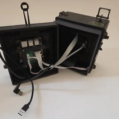 Innenaufnahme einer umgebauten Graflex Speed Graphic 4x5 Kamera. Das hintere Fach ist geöffnet und zeigt einen eingebauten Raspberry Pi 4, der über weiße Flachbandkabel mit einem Digitalsensor im vorderen Teil verbunden ist. Schwarze USB-Kabel für den Anschluss an die Powerbank.