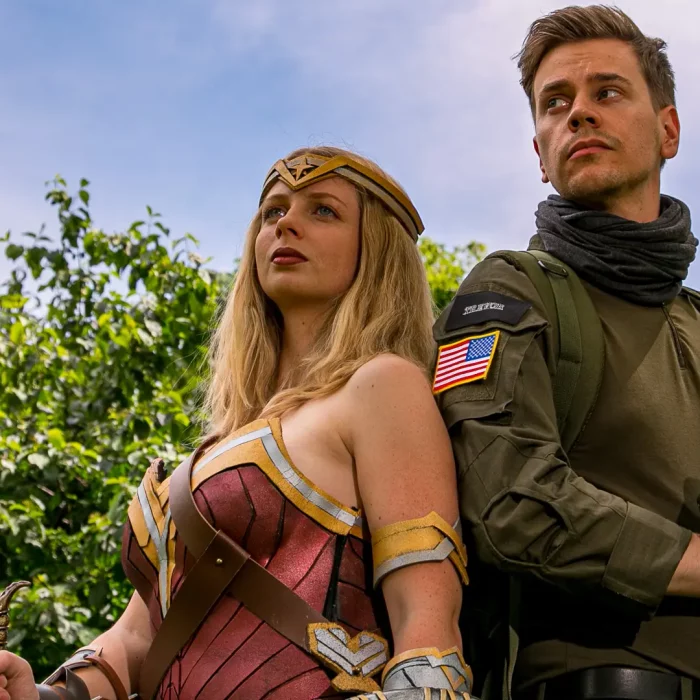 Frau im Wonder-Woman-Kostüm und Mann in Militär-Uniform, beide posieren vor grünem Laub im Freien